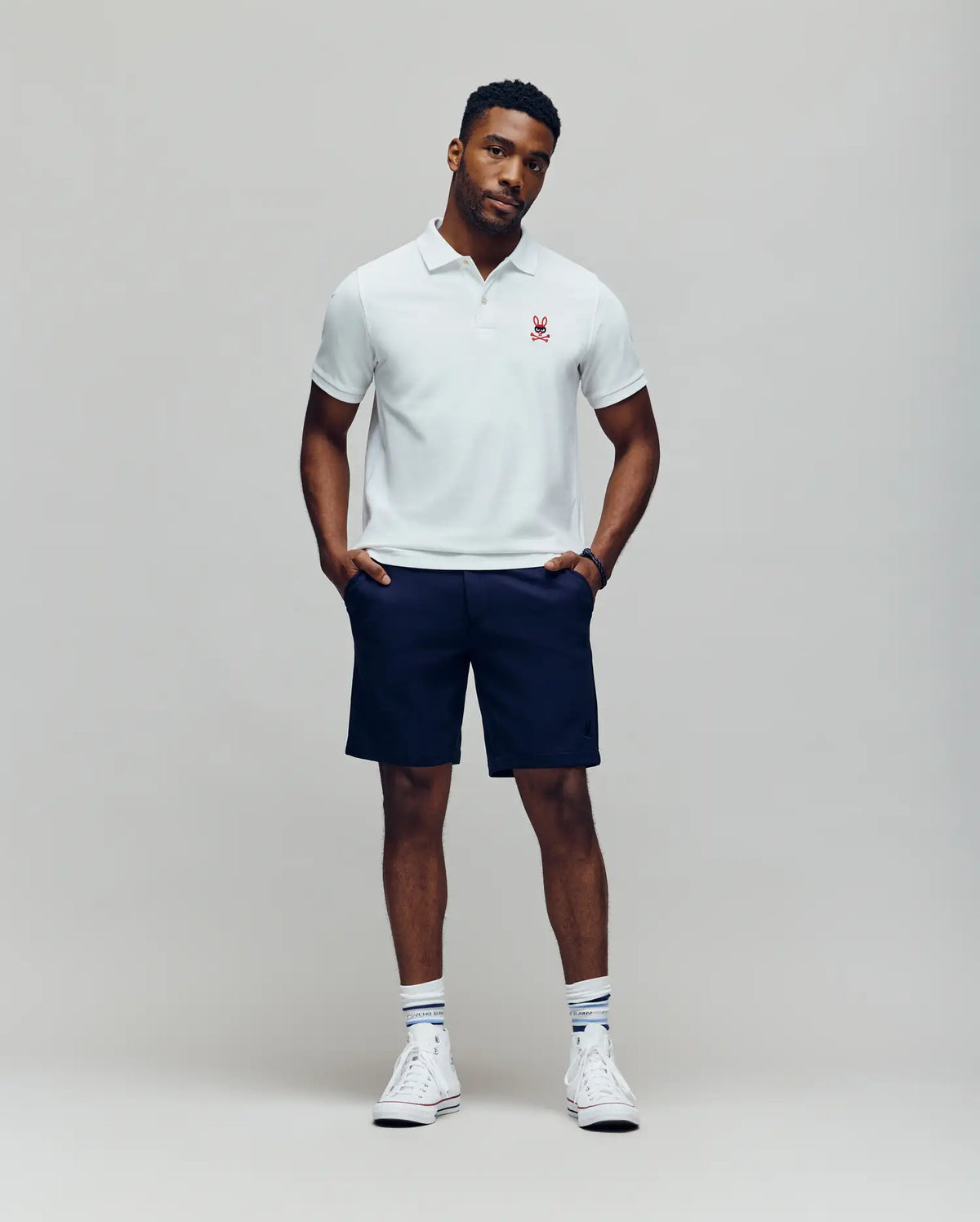 A man stands against a plain light gray background wearing a Psycho Bunny MENS MISCHIEF BUNNY PIQUE POLO - MT0100025 in white, paired with navy shorts, white socks, and white sneakers. His hands are in his pockets and his expression is neutral.