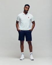 A man stands against a plain light gray background wearing a Psycho Bunny MENS MISCHIEF BUNNY PIQUE POLO - MT0100025 in white, paired with navy shorts, white socks, and white sneakers. His hands are in his pockets and his expression is neutral.