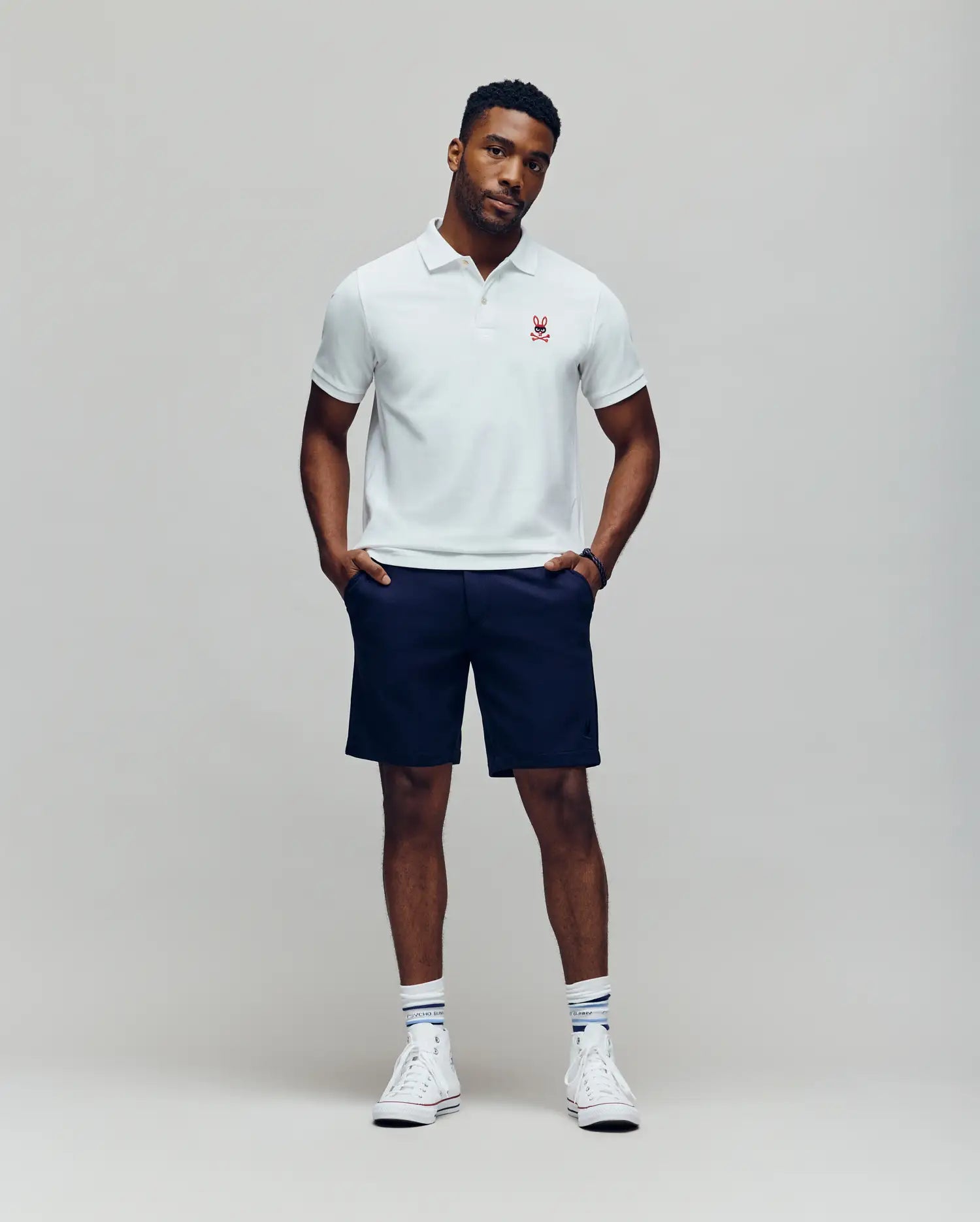 A man stands against a plain light gray background wearing a Psycho Bunny MENS MISCHIEF BUNNY PIQUE POLO - MT0100025 in white, paired with navy shorts, white socks, and white sneakers. His hands are in his pockets and his expression is neutral.