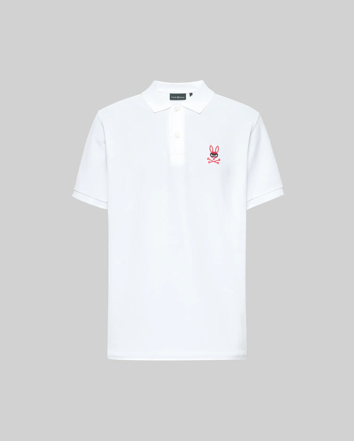 The Psycho Bunny MENS MISCHIEF BUNNY PIQUE POLO (MT0100025) is a white short-sleeve polo with a collar, three mother-of-pearl buttons, and a small pink bunny logo with crossbones on the left chest. Background is plain light gray.