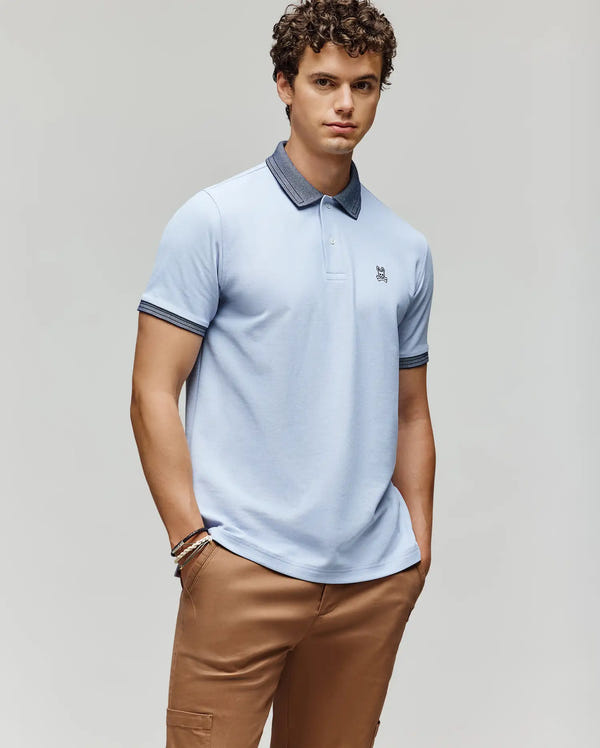 A young man with curly hair wears the Psycho Bunny MENS MURPHY PIQUE POLO SHIRT - MT0100026 in light blue, featuring an embroidered Bunny logo and contrast collar, paired with tan pants, standing against a plain light background.