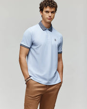 A young man with curly hair wears the Psycho Bunny MENS MURPHY PIQUE POLO SHIRT - MT0100026 in light blue, featuring an embroidered Bunny logo and contrast collar, paired with tan pants, standing against a plain light background.