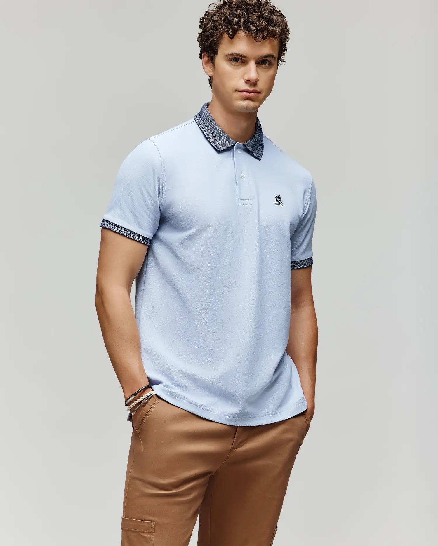 A young man with curly hair wears the Psycho Bunny MENS MURPHY PIQUE POLO SHIRT - MT0100026 in light blue, featuring an embroidered Bunny logo and contrast collar, paired with tan pants, standing against a plain light background.