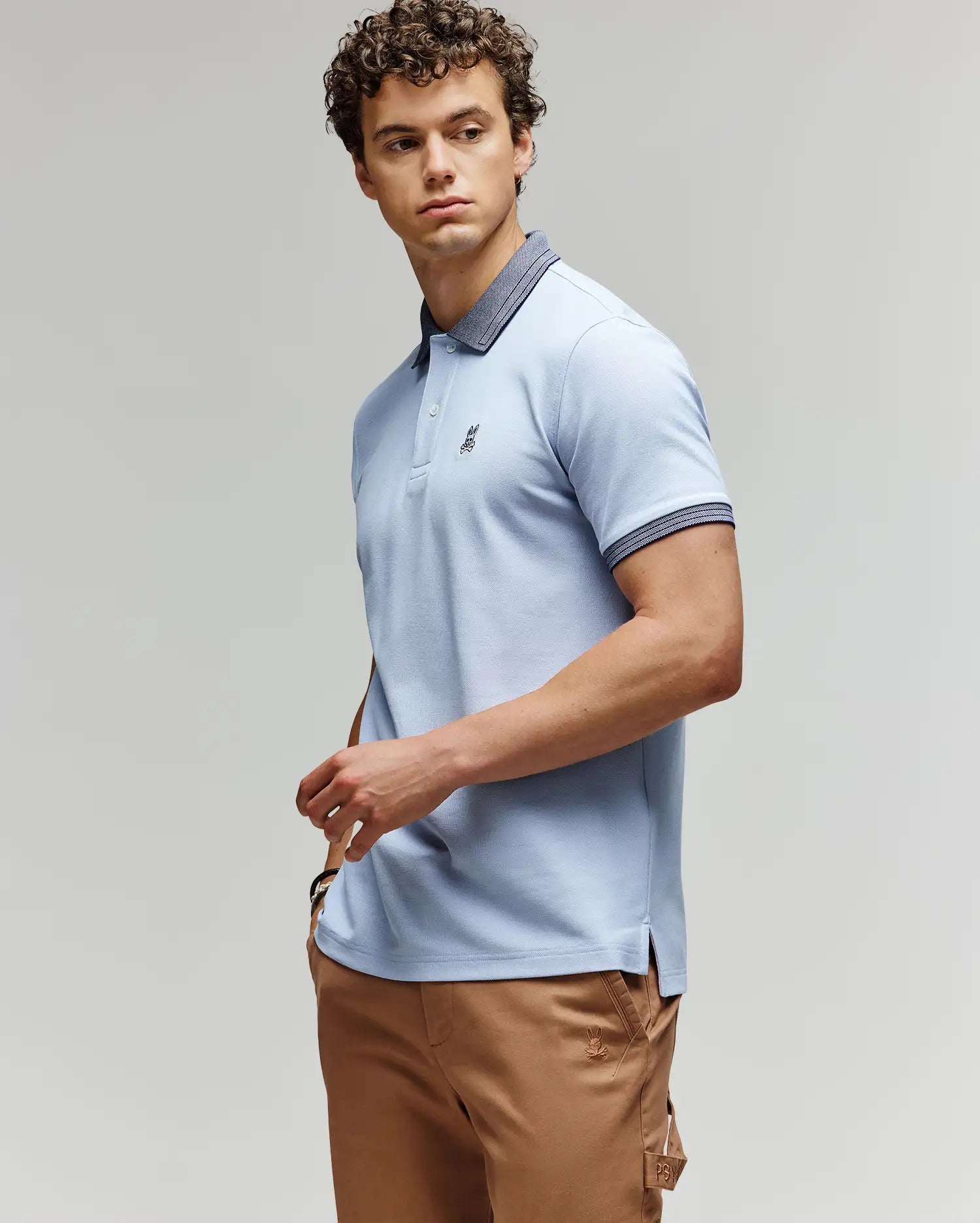 A man with curly hair wears Psycho Bunny’s MENS MURPHY PIQUE POLO SHIRT (MT0100026) in light blue, featuring an embroidered Bunny logo and contrast collar, paired with beige pants, standing against a plain light background.