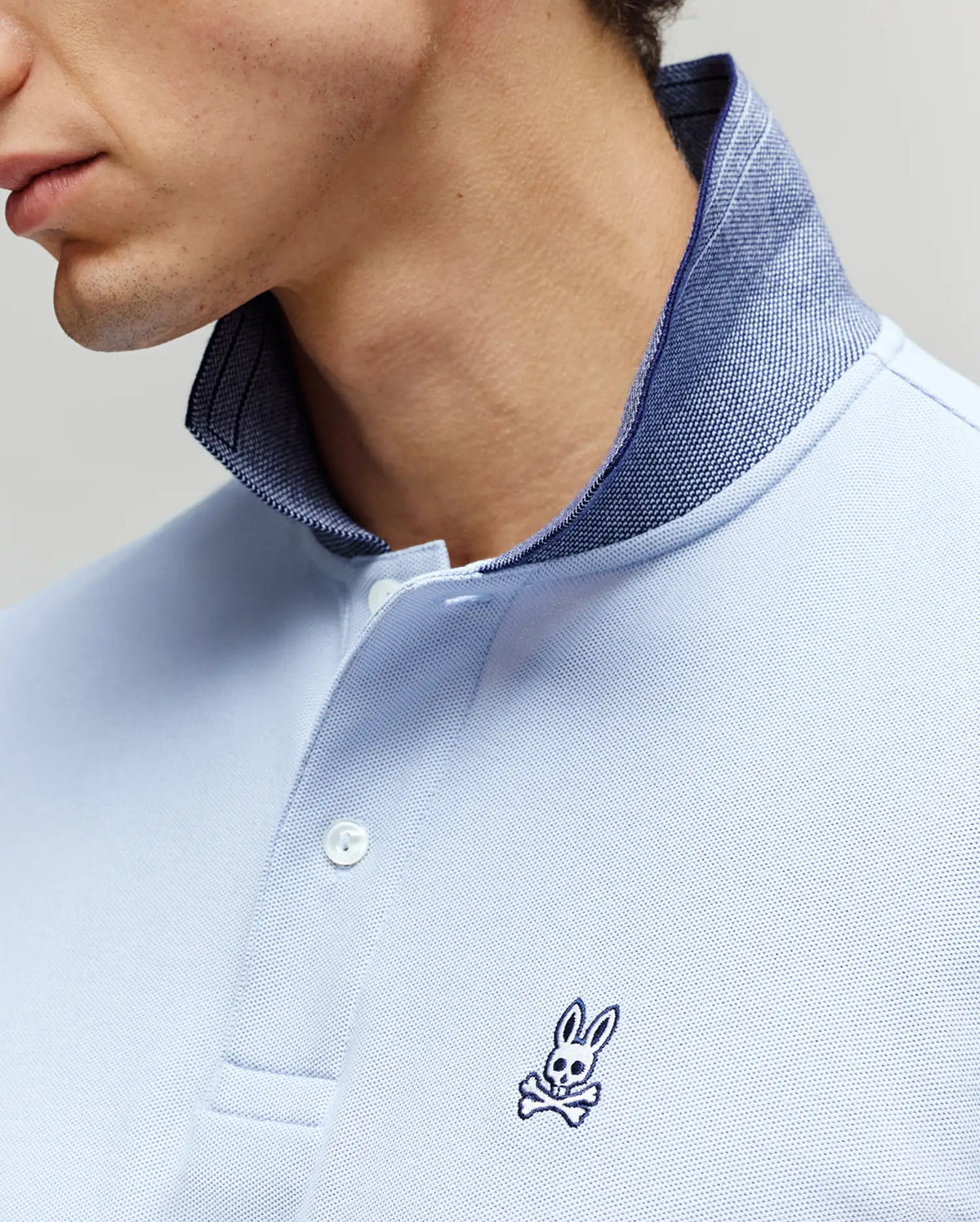 A close-up of a man wearing the Psycho Bunny MENS MURPHY PIQUE POLO SHIRT (MT0100026) in light blue, featuring a contrast collar and small embroidered Bunny logo on the chest. Only the lower half of his face and neck are visible.