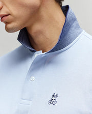 A close-up of a man wearing the Psycho Bunny MENS MURPHY PIQUE POLO SHIRT (MT0100026) in light blue, featuring a contrast collar and small embroidered Bunny logo on the chest. Only the lower half of his face and neck are visible.