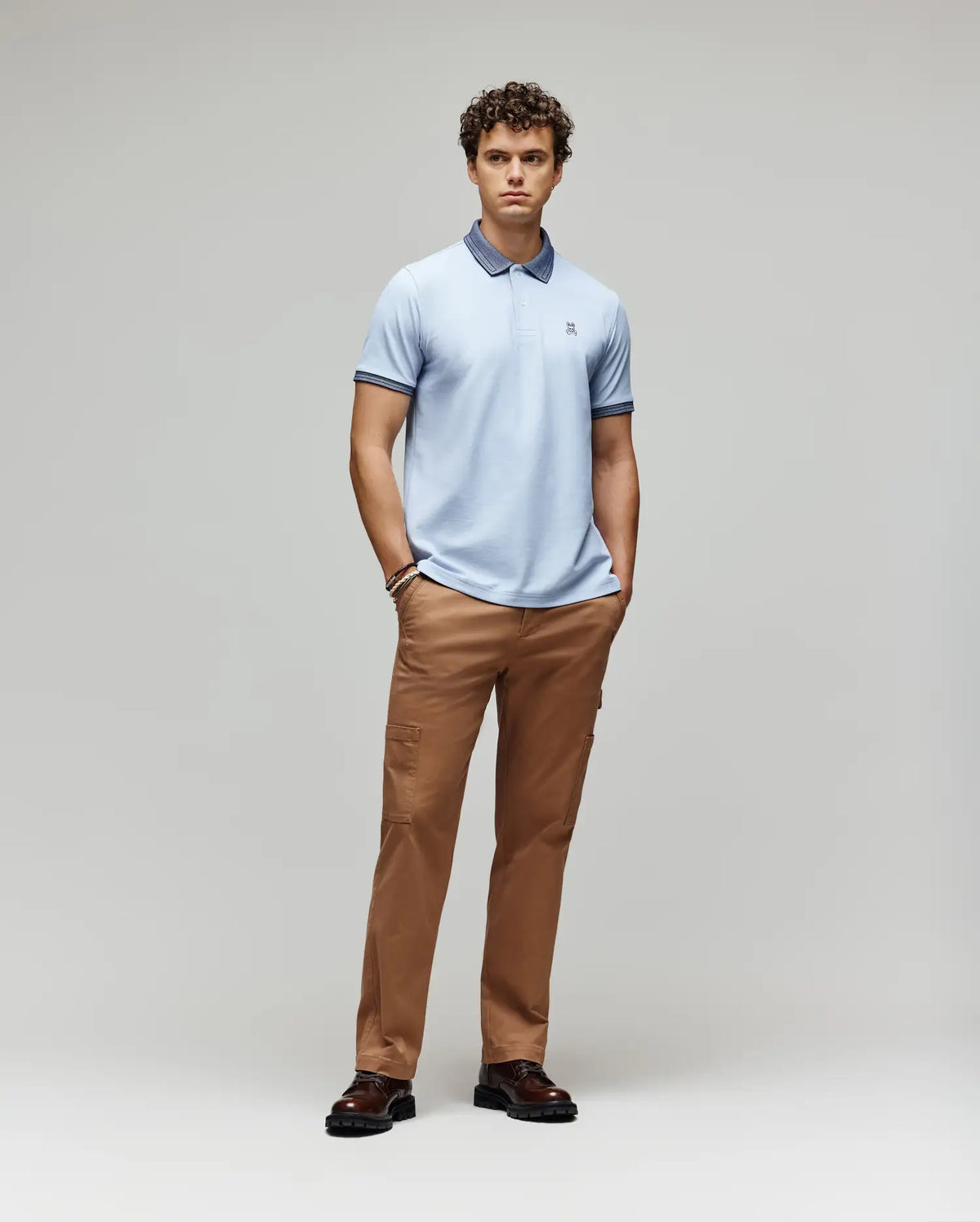 A man stands against a light gray background, wearing the Psycho Bunny MENS MURPHY PIQUE POLO SHIRT (MT0100026) in light blue with an embroidered Bunny logo, tan pants, and dark brown shoes, hands in pockets and facing forward neutrally.