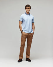 A man stands against a light gray background, wearing the Psycho Bunny MENS MURPHY PIQUE POLO SHIRT (MT0100026) in light blue with an embroidered Bunny logo, tan pants, and dark brown shoes, hands in pockets and facing forward neutrally.