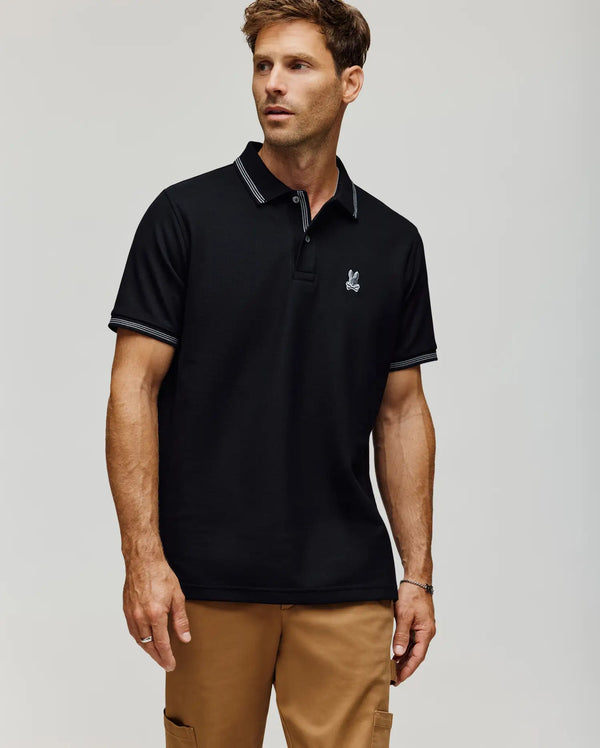A man stands against a plain light background wearing tan pants and the Psycho Bunny MENS HART PIQUE POLO SHIRT (MT0100032), a black Pima cotton polo featuring a small embroidered bunny logo on the chest.