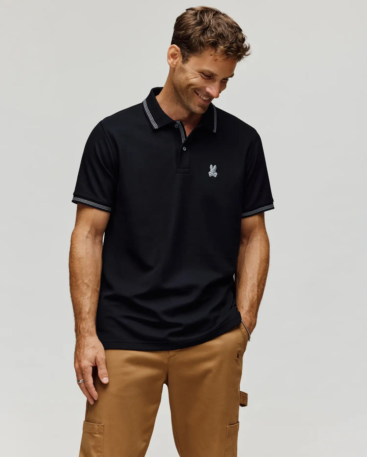 A man wearing the Psycho Bunny MENS HART PIQUE POLO SHIRT (MT0100032) in black, featuring a small embroidered logo, and tan cargo pants stands against a plain light background, smiling with his hands in his pockets.