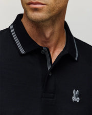 Close-up of a man in the Psycho Bunny MENS HART PIQUE POLO SHIRT (MT0100032), featuring black Pima cotton, white trim, and an embroidered rabbit with crossbones logo. His neutral expression highlights effortless menswear style.