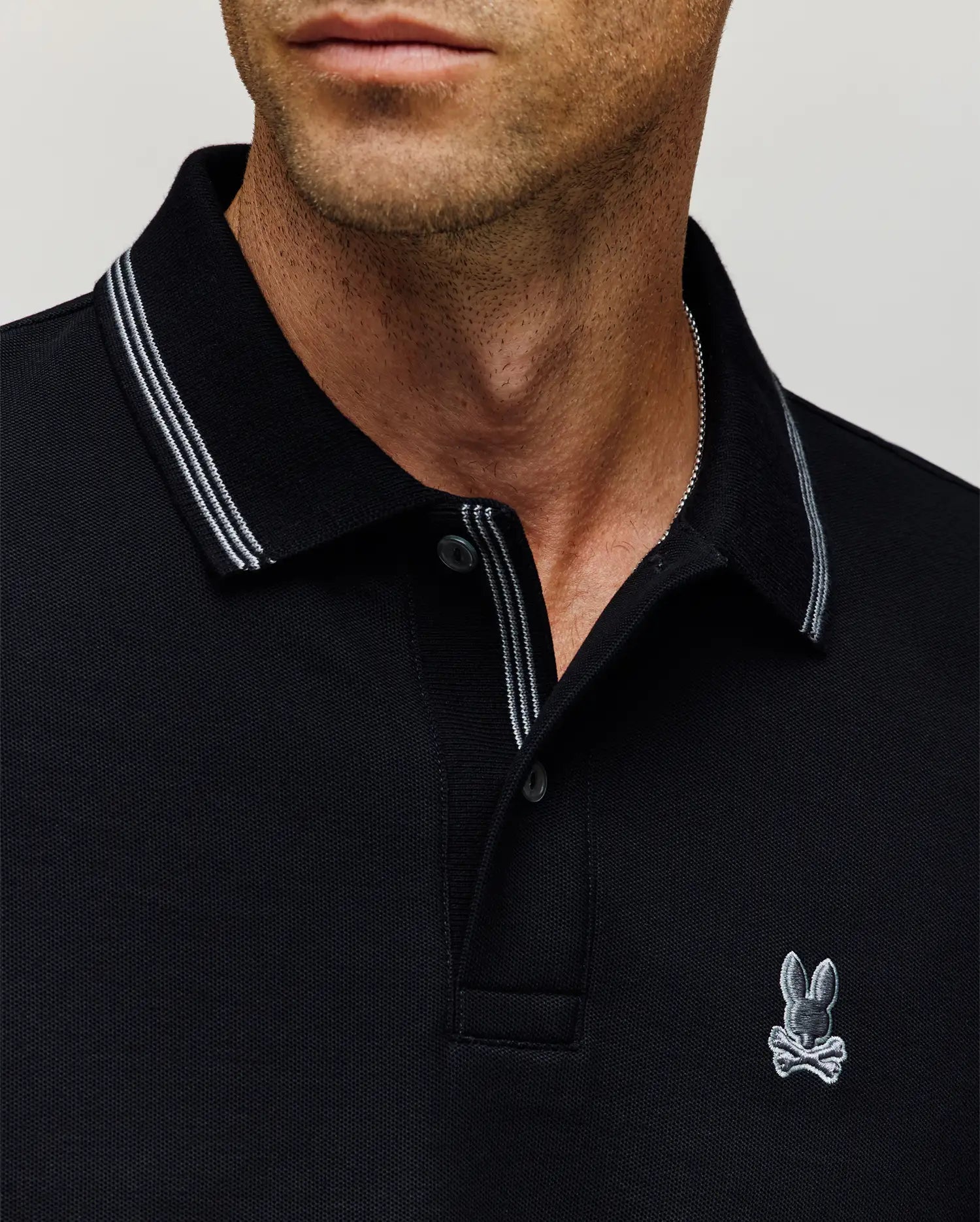 Close-up of a man in the Psycho Bunny MENS HART PIQUE POLO SHIRT (MT0100032), featuring black Pima cotton, white trim, and an embroidered rabbit with crossbones logo. His neutral expression highlights effortless menswear style.
