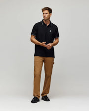 A man with short brown hair and a neutral expression wears a Psycho Bunny MENS HART PIQUE POLO SHIRT (MT0100032) in black, paired with brown cargo pants and black shoes, standing against a plain light gray background.