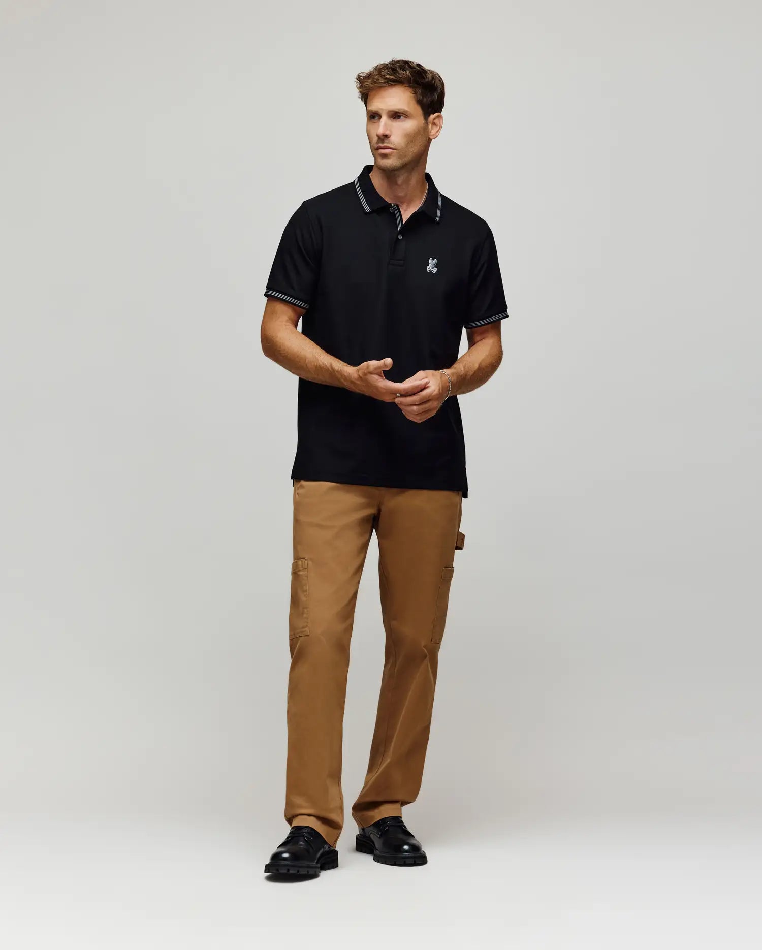 A man with short brown hair and a neutral expression wears a Psycho Bunny MENS HART PIQUE POLO SHIRT (MT0100032) in black, paired with brown cargo pants and black shoes, standing against a plain light gray background.