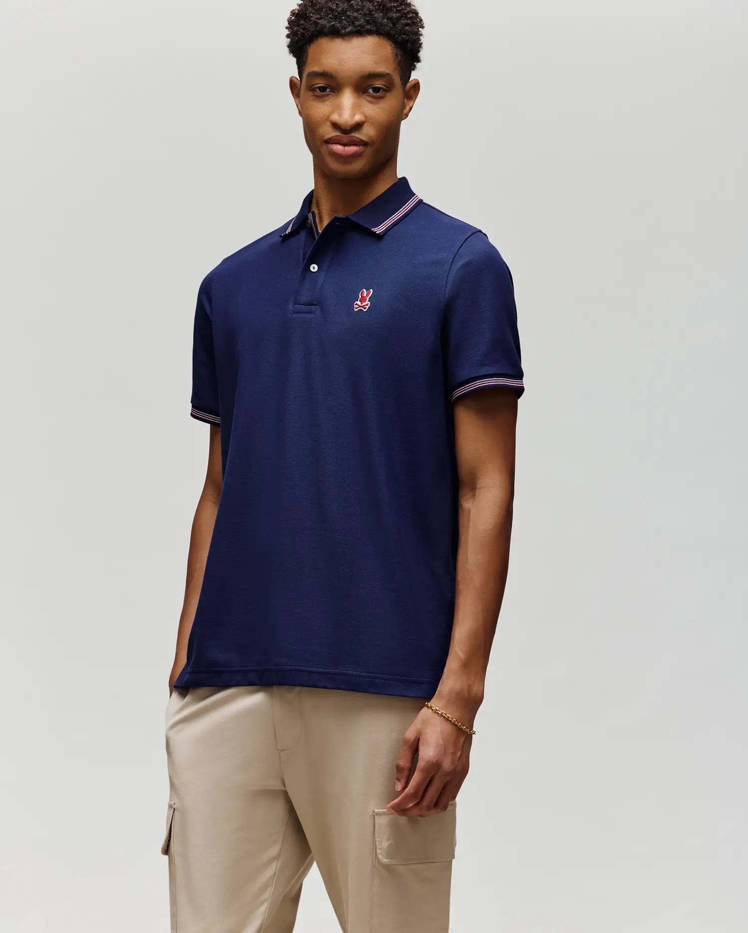 A man with short curly hair and a relaxed expression wears the Psycho Bunny MENS HART PIQUE POLO SHIRT - MT0100032 in navy blue, featuring a small red and white chest logo, paired with light beige cargo pants against a plain background.