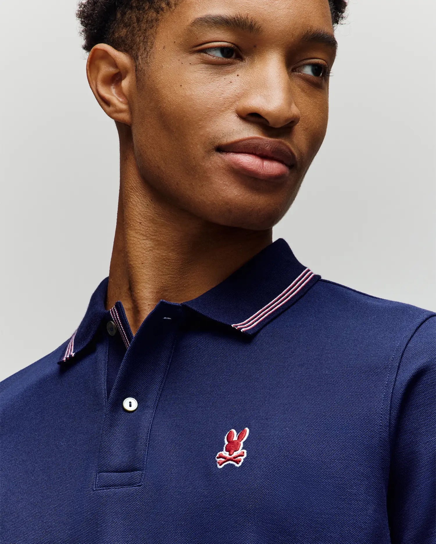 A man wears the Psycho Bunny MENS HART PIQUE POLO SHIRT (MT0100032) in navy, featuring a white and red striped collar and red bunny logo, modern fit, looking to the side against a neutral background.