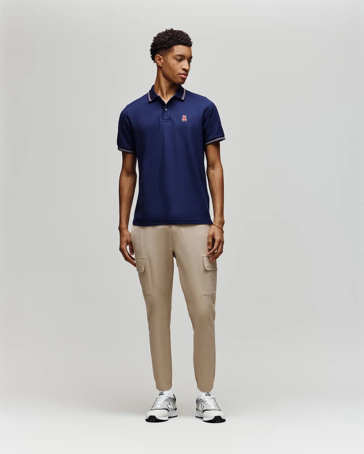 A man stands against a plain background wearing a Psycho Bunny MENS HART PIQUE POLO SHIRT - MT0100032 in navy, paired with beige cargo pants and white sneakers. He faces slightly to the side with his hands relaxed at his sides.