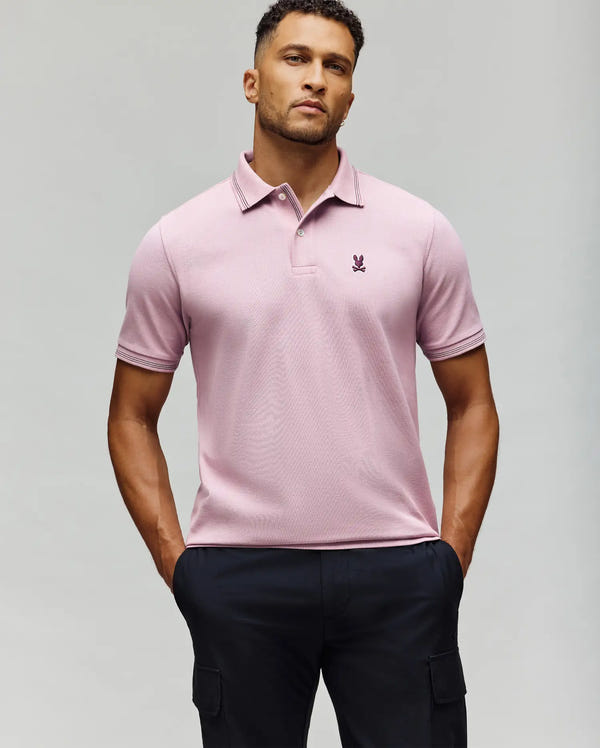 A man stands with hands in pockets, wearing the Psycho Bunny MENS HART PIQUE POLO SHIRT - MT0100032 in light pink and dark cargo pants, set against a plain light gray background.