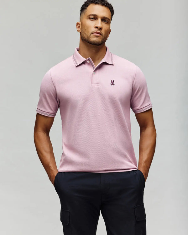 A man stands with hands in pockets, wearing the Psycho Bunny MENS HART PIQUE POLO SHIRT - MT0100032 in light pink and dark cargo pants, set against a plain light gray background.