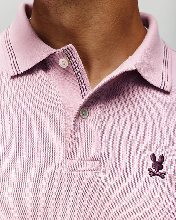 Close-up of the Psycho Bunny MENS HART PIQUE POLO SHIRT (MT0100032): a light pink Pima cotton polo with dark collar and button trim, featuring a small embroidered bunny head with crossbones logo on the left chest.