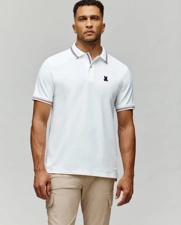 A man wears the Psycho Bunny MENS HART PIQUE POLO SHIRT (MT0100032) in white with a striped collar and sleeve edges, paired with light beige pants, standing against a plain grey background for a timeless menswear look.