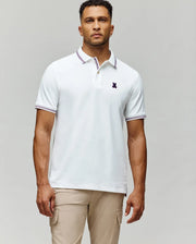 A man wears the Psycho Bunny MENS HART PIQUE POLO SHIRT (MT0100032) in white with a striped collar and sleeve edges, paired with light beige pants, standing against a plain grey background for a timeless menswear look.
