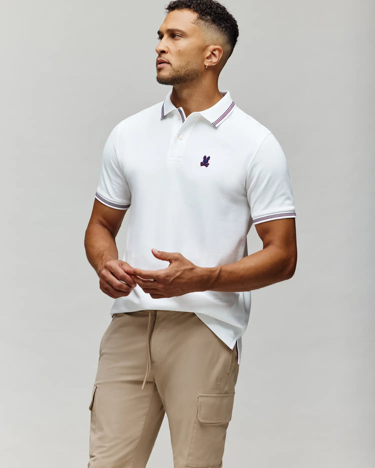 A man stands against a neutral background in Psycho Bunny's MENS HART PIQUE POLO SHIRT - MT0100032, featuring white Pima cotton and an embroidered purple logo, paired with beige cargo pants—a refined casual menswear look.
