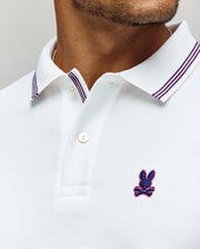A close-up of a man wearing the Psycho Bunny MENS HART PIQUE POLO SHIRT - MT0100032 in white, featuring a double-striped collar and the brand's signature blue and purple embroidered rabbit-with-crossbones logo on the chest.
