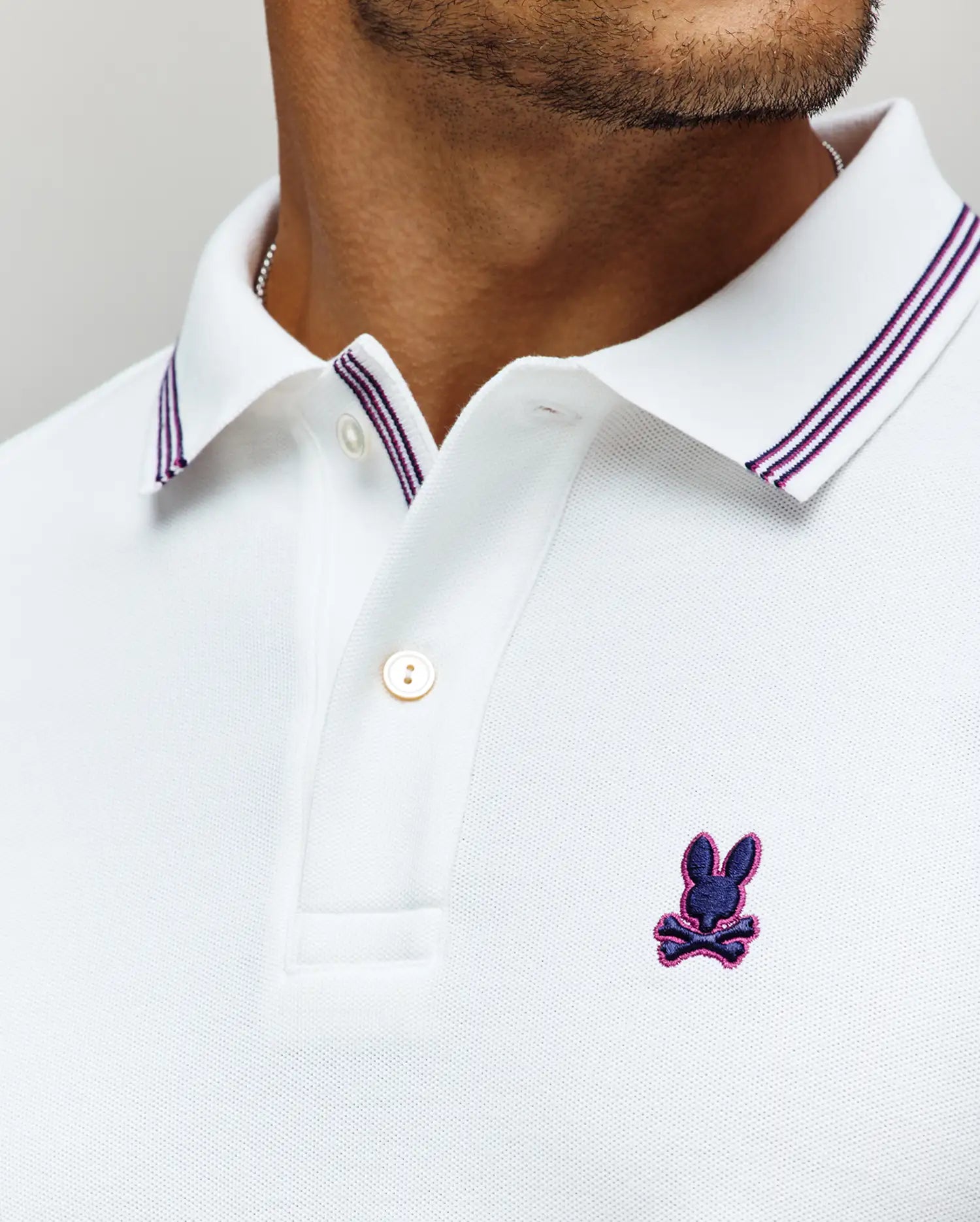 A close-up of a man wearing the Psycho Bunny MENS HART PIQUE POLO SHIRT - MT0100032 in white, featuring a double-striped collar and the brand's signature blue and purple embroidered rabbit-with-crossbones logo on the chest.