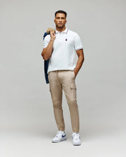 A man faces forward, wearing a white Psycho Bunny MENS HART PIQUE POLO SHIRT (MT0100032), beige cargo jogger pants, and white sneakers. He holds a dark jacket over his shoulder and keeps his other hand in his pocket.