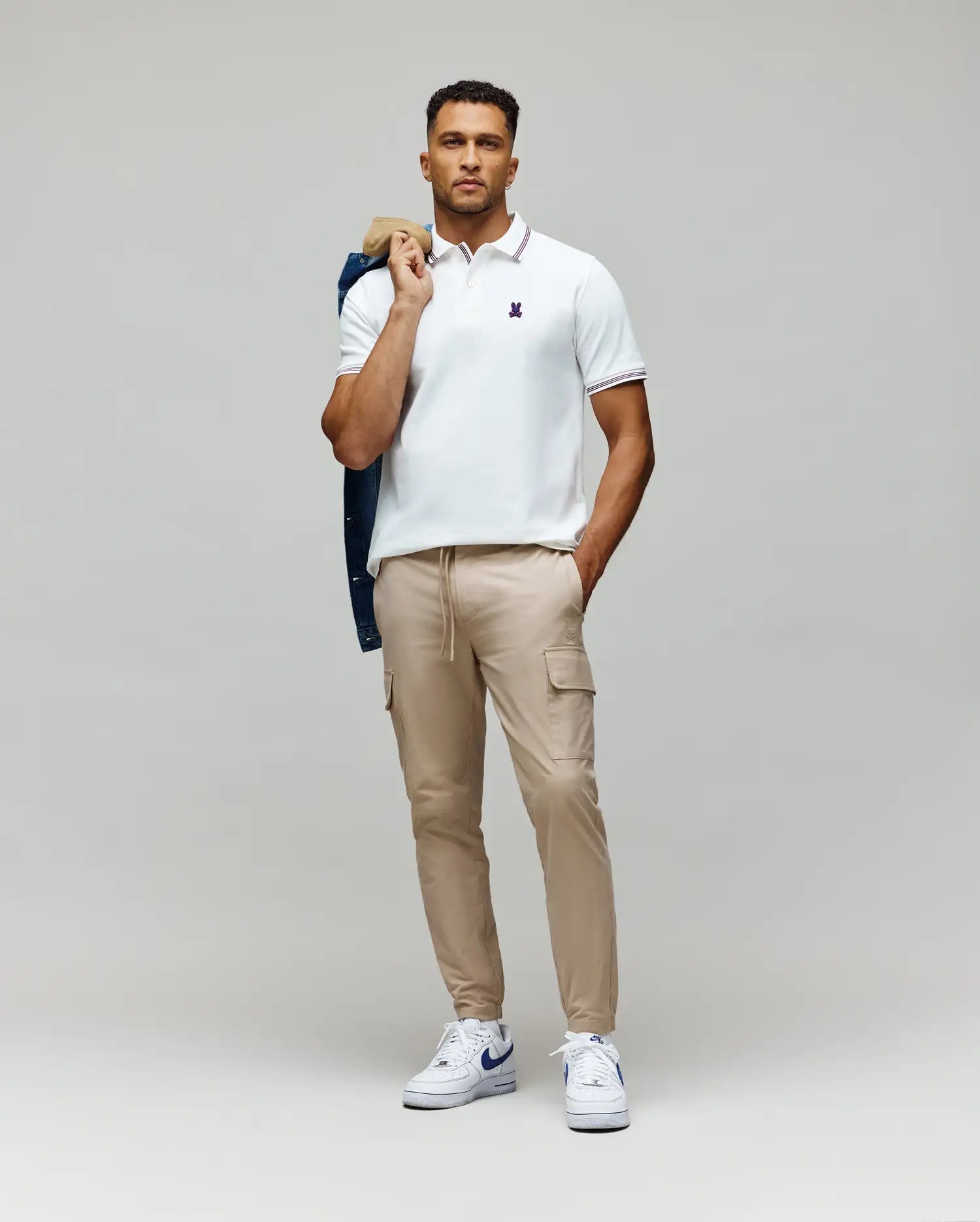 A man faces forward, wearing a white Psycho Bunny MENS HART PIQUE POLO SHIRT (MT0100032), beige cargo jogger pants, and white sneakers. He holds a dark jacket over his shoulder and keeps his other hand in his pocket.