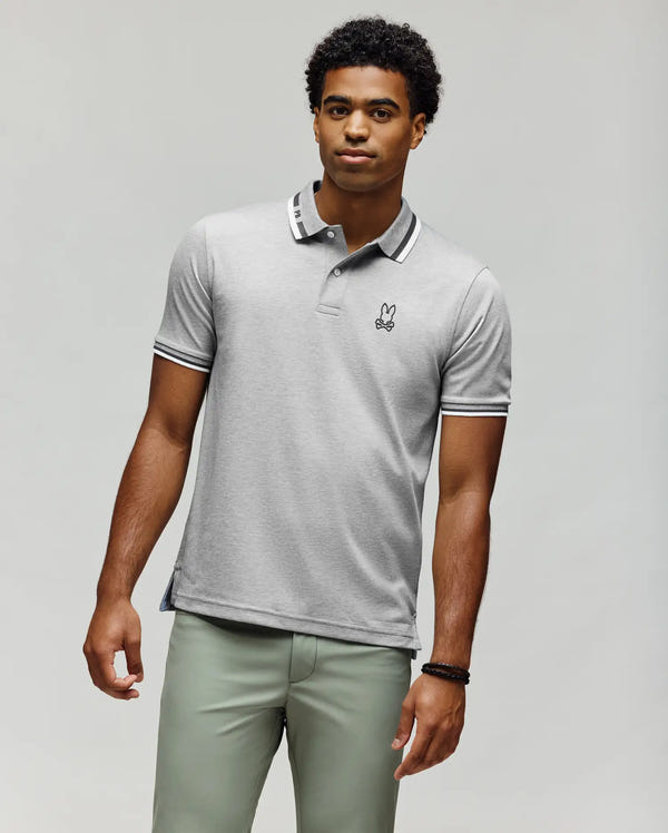 A man with short curly hair and a neutral expression wears Psycho Bunny’s slim fit MENS CHRIS PIQUE POLO SHIRT (MT0100038) with a small bunny logo, paired with light green pants against a plain background.