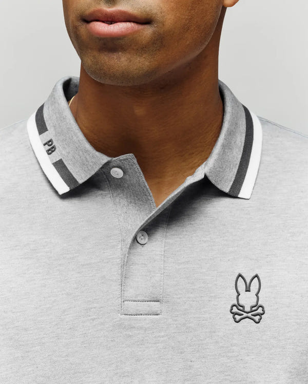 Close-up of a man in the Psycho Bunny MENS CHRIS PIQUE POLO SHIRT - MT0100038, light gray slim fit with dark and white striped collar, a bunny and crossbones chest logo, and "PB" stitched on the collar. His face is partially visible.