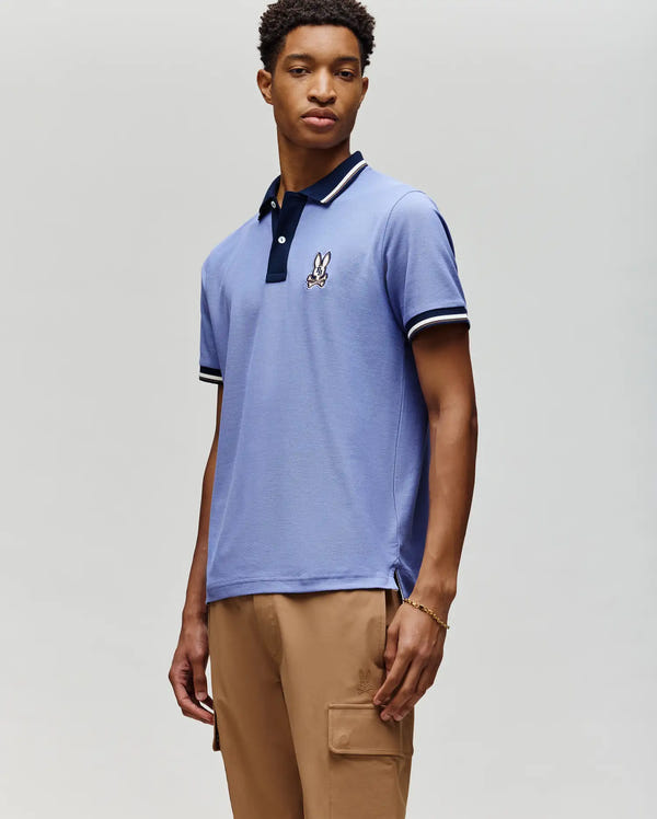 A young man stands against a plain background wearing the Psycho Bunny MENS WADE PIQUE POLO SHIRT (MT0100044) in light blue with an embroidered bunny logo, paired with khaki cargo pants and a gold bracelet, gazing slightly to the side.