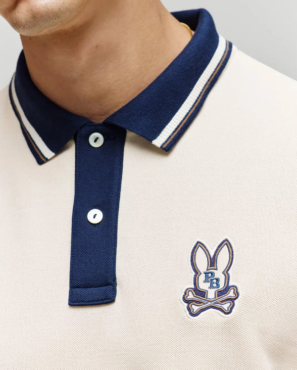 Close-up of a person wearing the Psycho Bunny MENS WADE PIQUE POLO SHIRT (MT0100044) in beige, featuring a navy blue collar and buttons, with an embroidered bunny and crossed bones logo on the left chest.