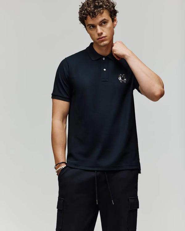 A young man with curly hair wears a Psycho Bunny MENS AUGUSTE PIQUE POLO SHIRT - MT0100051 and black cargo pants, standing against a plain light background and looking at the camera with a neutral expression.