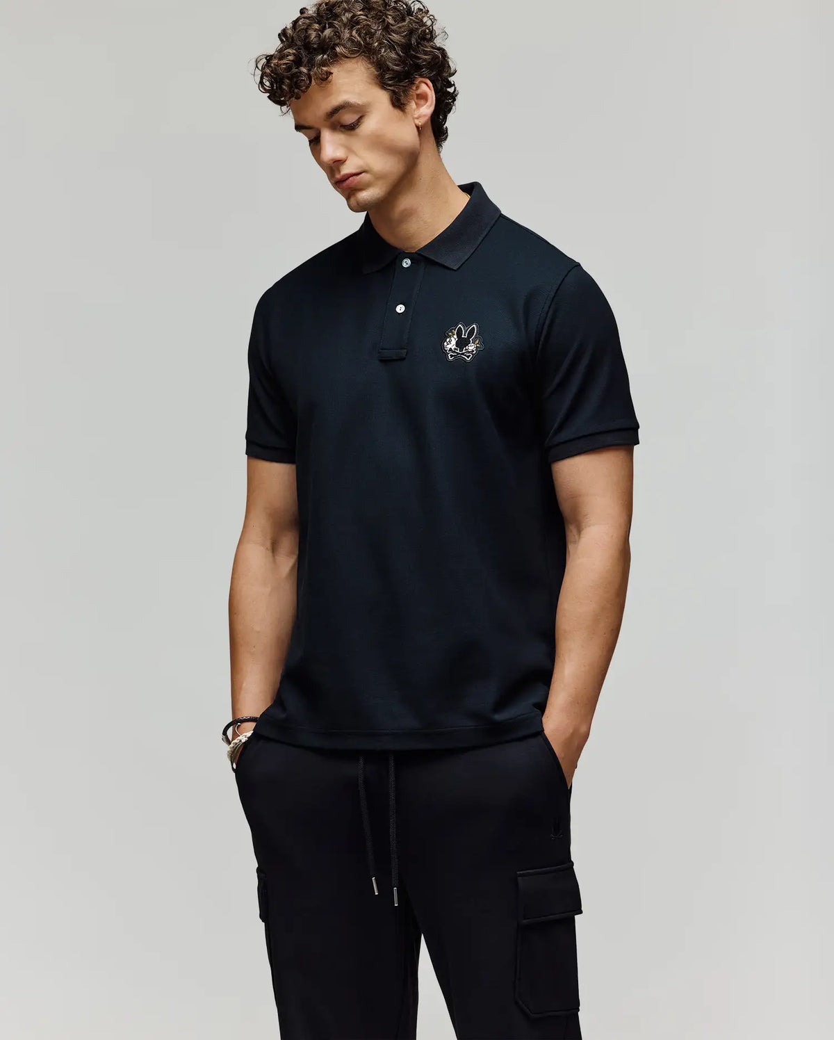 A man with curly hair stands with his hands in his pockets, wearing Psycho Bunny’s MENS AUGUSTE PIQUE POLO SHIRT - MT0100051 in black and black cargo pants against a plain background.