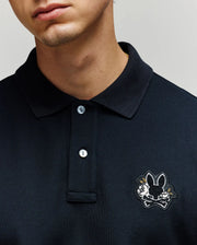 A man wears a Psycho Bunny MENS AUGUSTE PIQUE POLO SHIRT (MT0100051) in black, featuring an embroidered black bunny head with crossed bones patch on the left chest. Only his lower face and upper torso are visible.