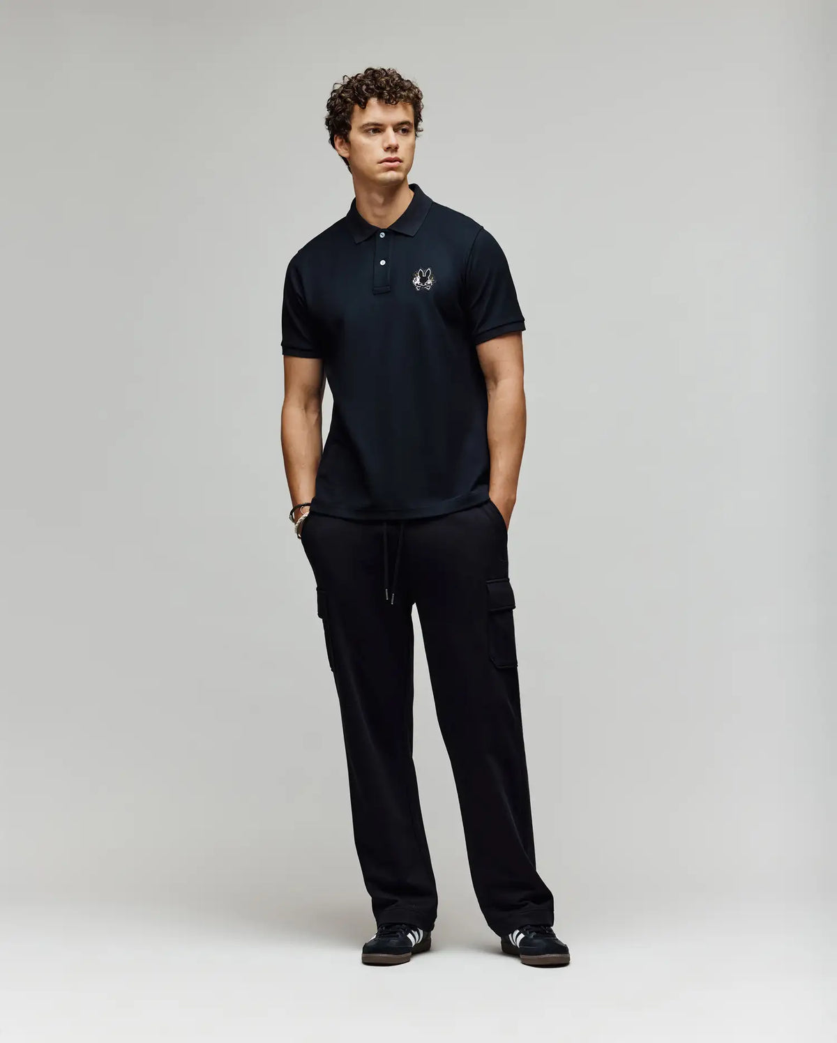 A man with curly hair stands against a plain light background, wearing the Psycho Bunny MENS AUGUSTE PIQUE POLO SHIRT - MT0100051 in black, paired with black cargo pants and black sneakers with white stripes, hands in pockets for a classic Americana vibe.
