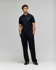 A man with curly hair stands against a plain light background, wearing the Psycho Bunny MENS AUGUSTE PIQUE POLO SHIRT - MT0100051 in black, paired with black cargo pants and black sneakers with white stripes, hands in pockets for a classic Americana vibe.