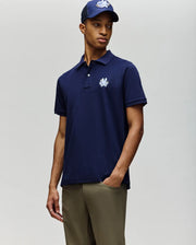 A man stands with one hand in his pocket, wearing the Psycho Bunny MENS AUGUSTE PIQUE POLO SHIRT - MT0100051 in navy blue, a matching navy baseball cap, and olive green pants against a plain background.