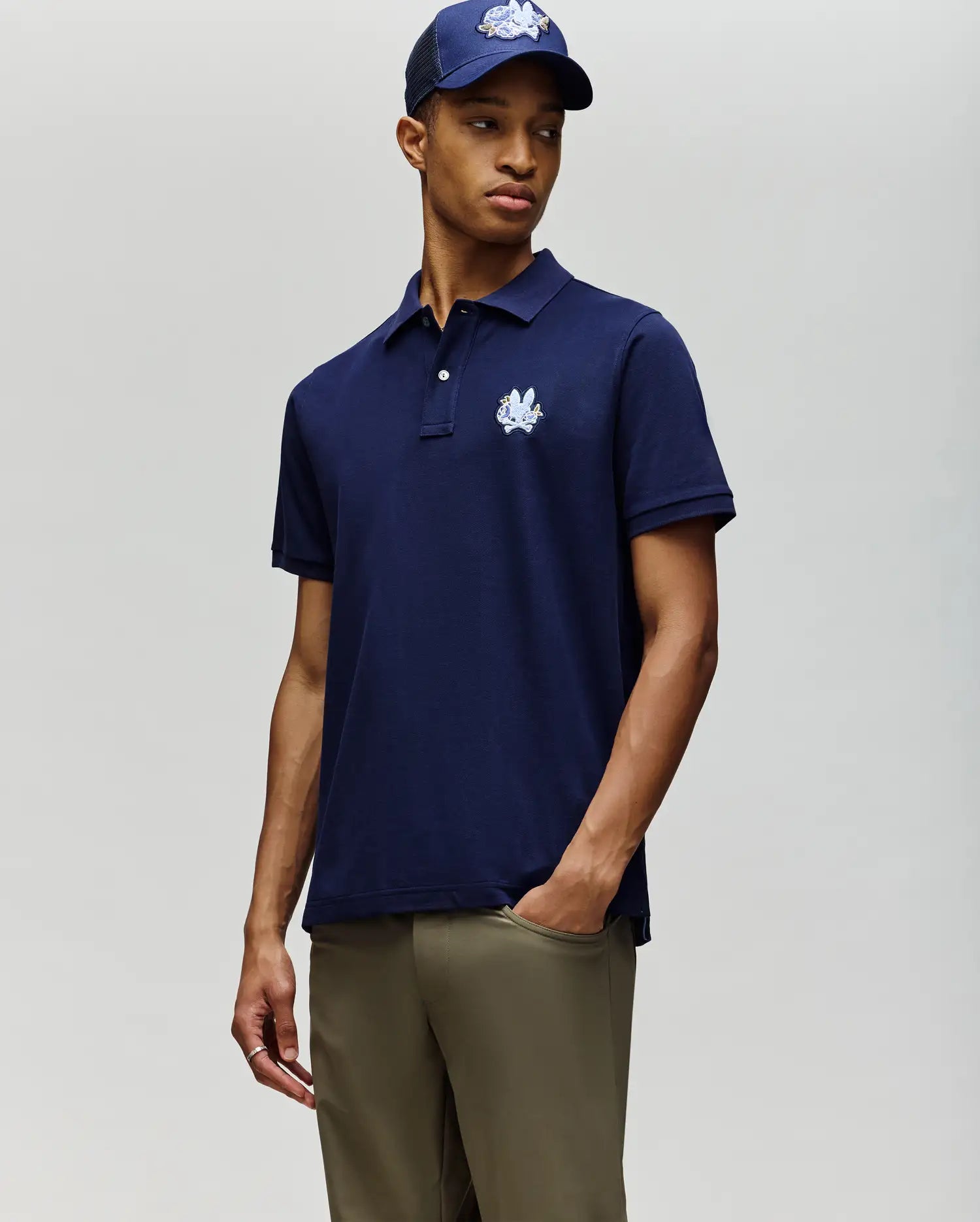 A man stands with one hand in his pocket, wearing the Psycho Bunny MENS AUGUSTE PIQUE POLO SHIRT - MT0100051 in navy blue, a matching navy baseball cap, and olive green pants against a plain background.