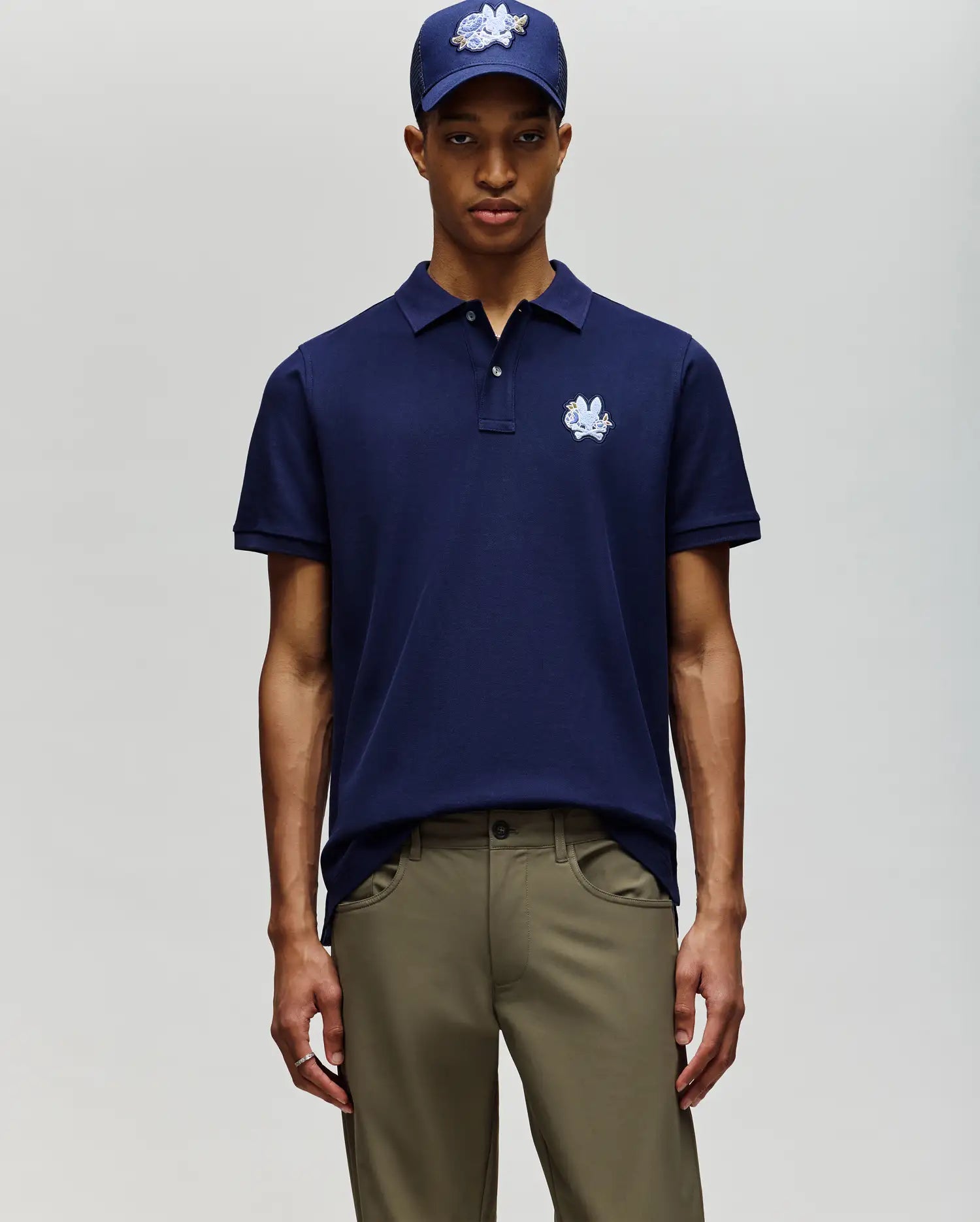 A man stands facing forward against a plain light background, wearing the Psycho Bunny MENS AUGUSTE PIQUE POLO SHIRT (MT0100051) in navy blue, a matching embroidered cap, and olive green pants.