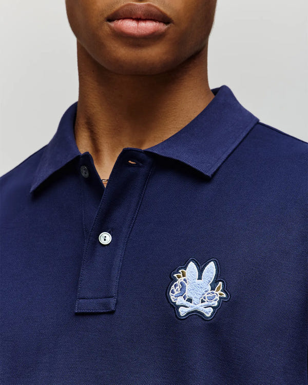 Close-up of a person in the Psycho Bunny MENS AUGUSTE PIQUE POLO SHIRT (MT0100051), dark blue Pima cotton with a collared design and an embroidered bunny and crossed bones patch on the left chest. The face is partially visible.