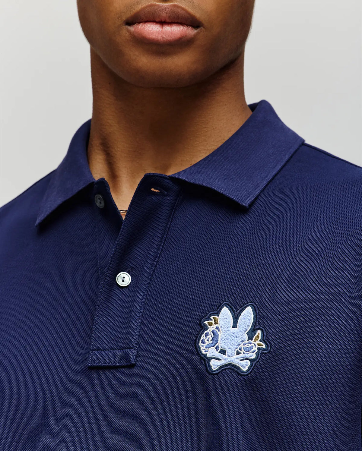 Close-up of a person in the Psycho Bunny MENS AUGUSTE PIQUE POLO SHIRT (MT0100051), dark blue Pima cotton with a collared design and an embroidered bunny and crossed bones patch on the left chest. The face is partially visible.