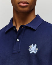 Close-up of a person in the Psycho Bunny MENS AUGUSTE PIQUE POLO SHIRT (MT0100051), dark blue Pima cotton with a collared design and an embroidered bunny and crossed bones patch on the left chest. The face is partially visible.