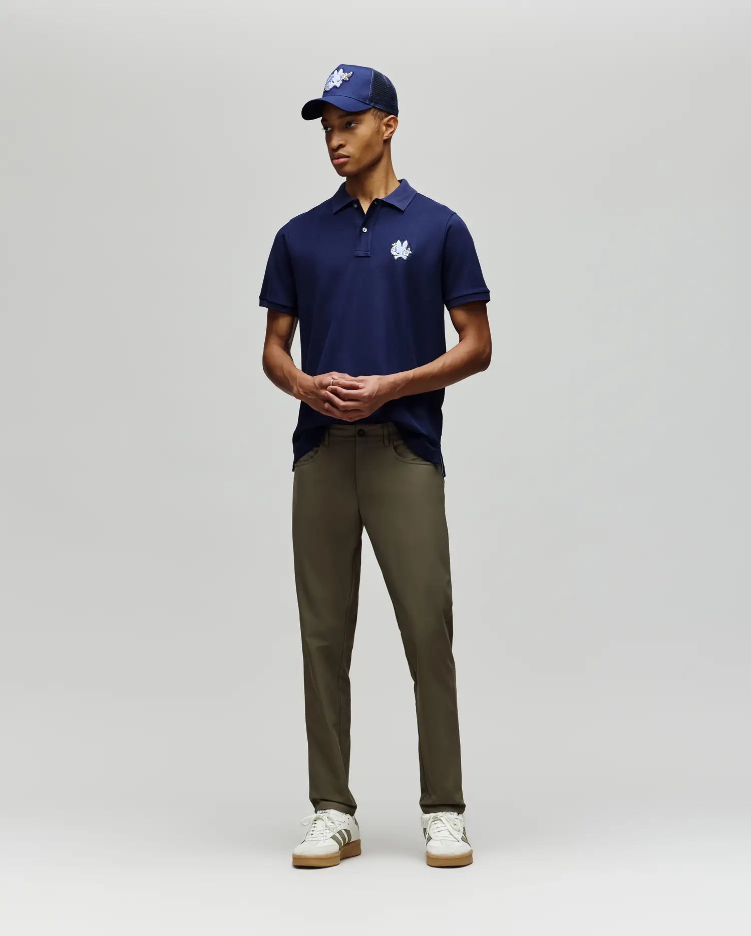 A man stands against a plain background wearing the Psycho Bunny MENS AUGUSTE PIQUE POLO SHIRT - MT0100051 in navy blue with a white logo, olive green pants, white sneakers, and a matching navy varsity cap with a white logo.