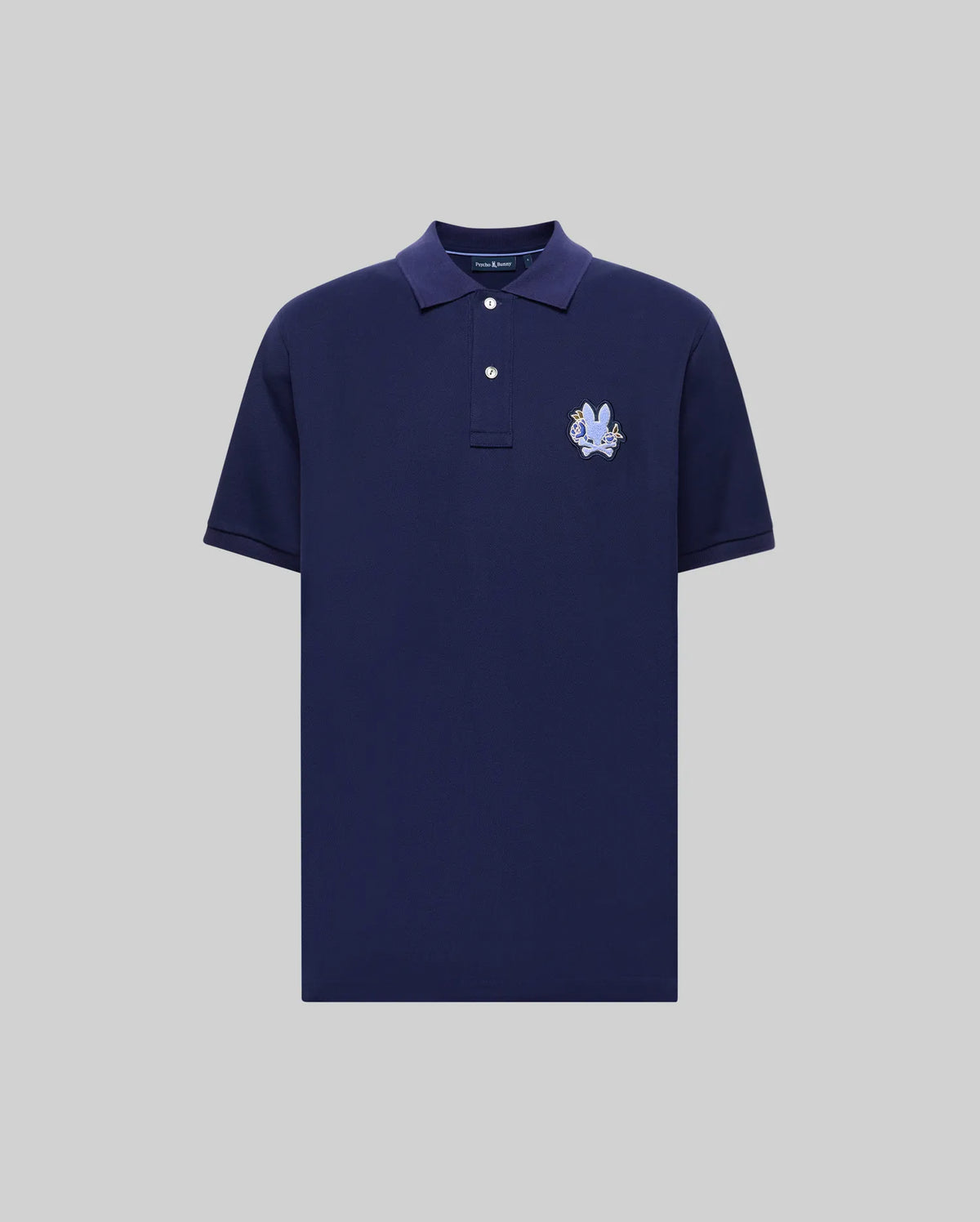The Psycho Bunny MENS AUGUSTE PIQUE POLO SHIRT (MT0100051) is a navy blue short-sleeve Pima cotton polo with a collar, two white buttons, and a light blue embroidered crest on the left chest, shown on a light gray background.