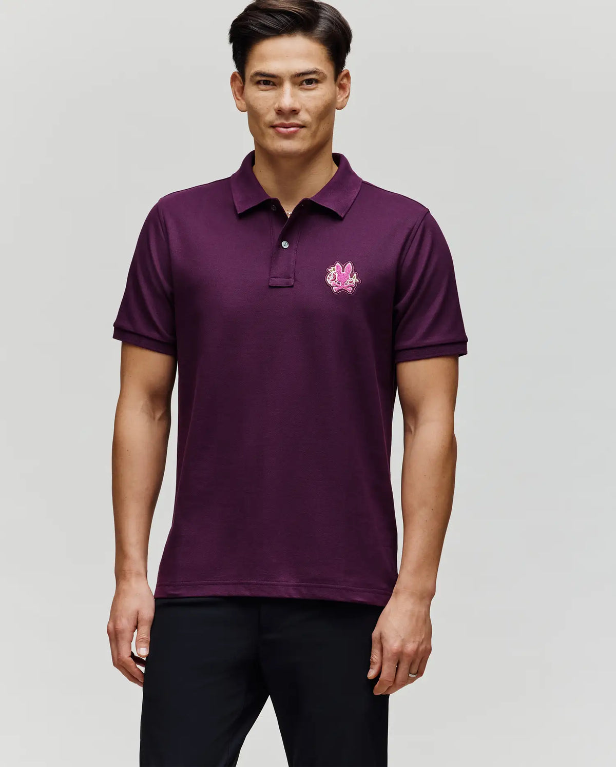 A man faces forward wearing Psycho Bunny's MENS AUGUSTE PIQUE POLO SHIRT - MT0100051, a short-sleeve dark purple pima cotton polo with an embroidered logo on the left chest, paired with black pants against a plain light grey background.