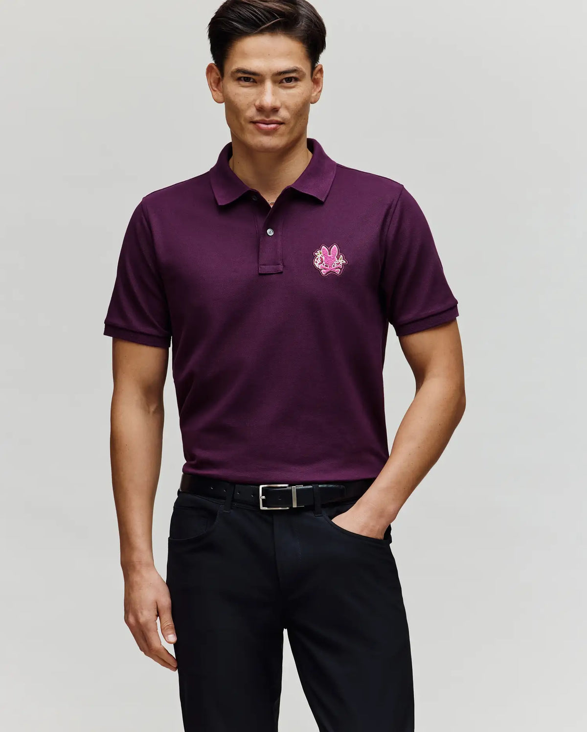 A man smiles with one hand in his pocket, wearing Psycho Bunny's MENS AUGUSTE PIQUE POLO SHIRT (MT0100051) in dark purple, featuring a small embroidered logo on the chest, paired with black pants and a black belt against a plain background.